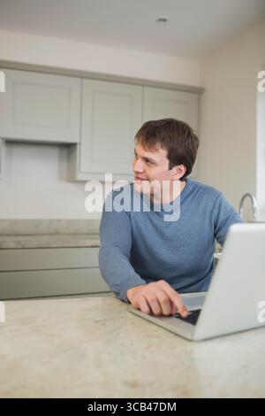 Mann, der an einem Laptop auf der Marmor-Kücheninsel arbeitet, während er links in Richtung graue Schränke und Wasserhahn blickte Stockfoto