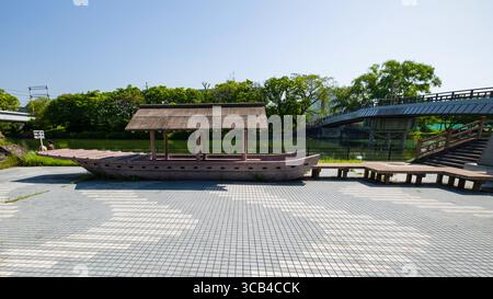 Auf dem Fushimi Minato Square befinden sich ein Jukoku-Boot-Modell und eine moderne Brücke unter einem klaren blauen Himmel. Üppiges Grün umgibt die Szene und schafft ein ruhiges atmos Stockfoto
