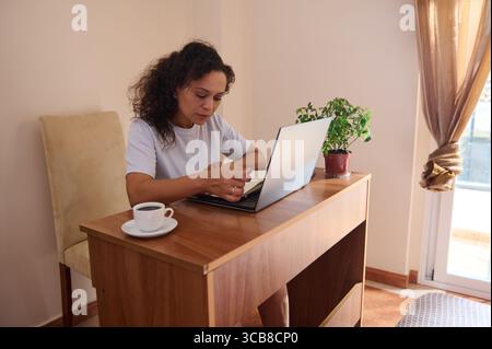 Eine Frau sitzt an einem hölzernen Schreibtisch in einem gemütlichen, sonnendurchfluteten Raum und konzentriert sich auf ihren Laptop. Eine Tasse Kaffee steht in der Nähe und vermittelt ein Gefühl von Produktivität und relaxa Stockfoto