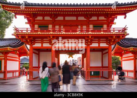 Ein leuchtendes rot-weißes Yasaka Jinja Nishiromon Tor, das mit goldenen Akzenten geschmückt ist, mit Besuchern, die durch die Stadt kommen. Die Szene zeigt architektonische Beau Stockfoto