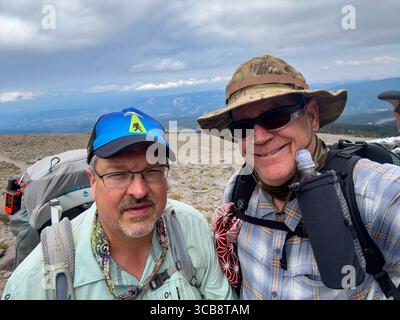 Zwei Freunde genießen ein Wanderabenteuer im Freien auf dem Mount Hood Timberline Trail, der Rucksäcke durch eine malerische Bergregion mit bewölktem Himmel führt Stockfoto
