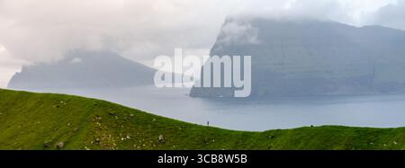 Klippen erheben sich dramatisch über das ruhige Wasser, während Nebel die Landschaft der Färöer bedeckt. Eine Einzelfigur steht im Vordergrund, Kallur Lighthouse, Kalsoy Island Stockfoto