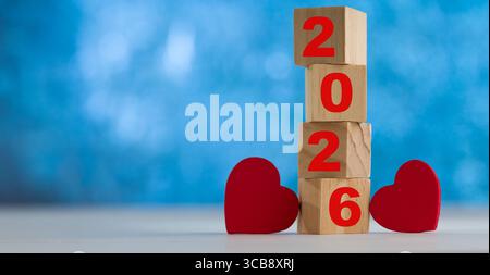 Das Wort „2026“ aus Holzblöcken und zwei roten Herzen auf blauem, verschwommenem Hintergrund symbolisiert Liebe, Hoffnung und das Thema Neujahr. Stockfoto