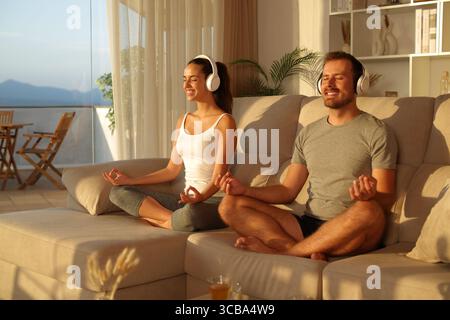 Glückliches Paar, das Yoga-Kurs von einem Kopfhörer aus hört und zu Hause bei Sonnenuntergang auf einem Sofa sitzt Stockfoto