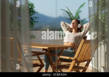 Frau, die sich entspannt auf einer Terrasse sitzt und Musik zu Hause hört, an einem sonnigen Tag Stockfoto