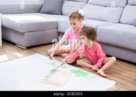 Zwei Schwestern sitzen auf dem Wohnzimmerboden und malen zusammen auf einem großen Blatt Papier mit bunten Farben und Pinseln. Stockfoto