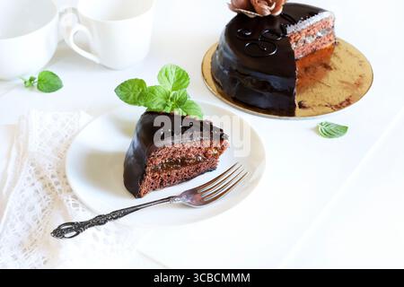 Sachertorte (Schokoladen-sacher-Kuchen) - Scheibe auf einem weißen Teller, frische Minzdekoration, ganzer Kuchen im Hintergrund. Traditionelles österreichisches Dessert. Stockfoto