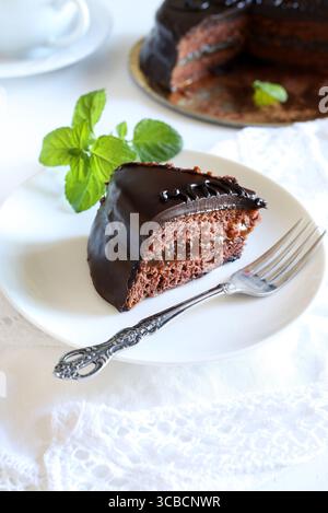 Sachertorte (Schokoladen-sacher-Kuchen) - Scheibe auf einem weißen Teller, frische Minzdekoration, ganzer Kuchen im Hintergrund. Traditionelles österreichisches Dessert. Dre Stockfoto