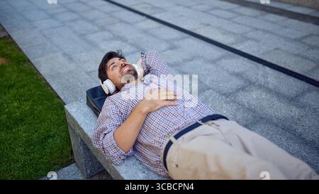 Lächelnder Mann mit Kopfhörern, der draußen auf einer Bank liegt Stockfoto
