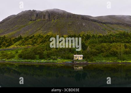 Kleines gelbes Haus, das sich in einem See mit grünen Bäumen und Bergen spiegelt Stockfoto