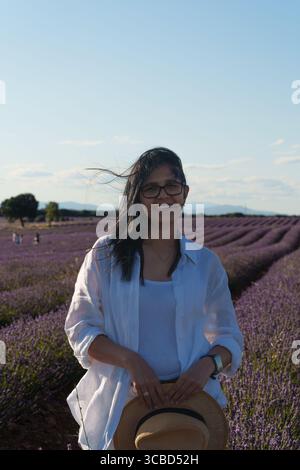 Eine Frau mit weißem Hemd und Strohhut steht auf einem Lavendelfeld. Sie lächelt und genießt die Landschaft Stockfoto