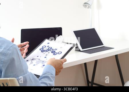 Mann in blauem Hemd mit Klemmbrett mit medizinischen Symbolen und Weltkarte, erklärt Daten, während er an einem modernen Schreibtisch mit digitalen Tablets in einem Gesundheitswesen sitzt Stockfoto