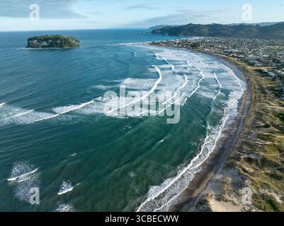 Aus der Vogelperspektive auf das türkisfarbene Meer trifft auf die sandigen Küsten in der Nähe einer kleinen Insel, mit der Stadt an der Küste, Whangamatā, Waikato Region, Neuseeland. Stockfoto