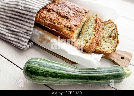 Rezept für Zucchini- und Käsebrot, ein Brotlaib auf einem hölzernen Schneidebrett, bedeckt mit einem Küchentuch, daneben ein ganzes Zucchini-Gemüse. Stockfoto