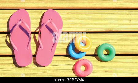 Leuchtend rosa Flipflops ruhen auf gelben Holzterrassen neben farbenfrohen Schwimmerspielzeugen. Die lebhafte Szene fängt einen sonnigen Tag ein, perfekt zum Entspannen Stockfoto