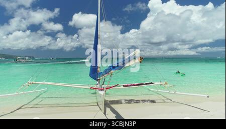 Türkisfarbenes tropisches Wasser, das sanft am Ufer des White Beach in Boracay, Philippinen, liegt mit einem traditionellen Paraw-Segelboot auf unberührtem Sand unter einem leuchtend blauen Himmel Stockfoto