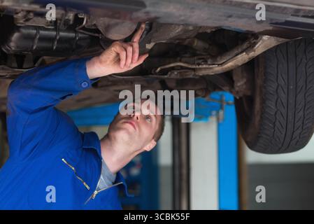 Männlicher Kfz-Techniker, der auf den Auspuff zeigt, untersuchte das Laufwerk auf dem Aufzug in der Werkstatt, Kopierraum. Mechaniker, Wartung, Inspektion, Werkstatt, Prec Stockfoto