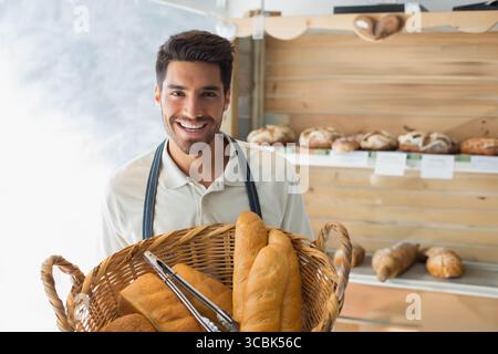 Erwachsener männlicher Bäcker in dunkler Schürze, der Brote mit einer Metallzange an der Backstube in den Korb legt. Handwerker, Bäckerei, handgefertigt, rustikal, frisch, Gourmet, in Stockfoto