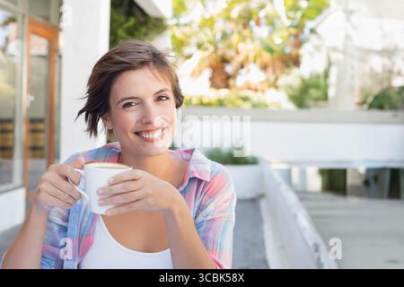 Frau in den Zwanzigern, die auf der Terrasse des Cafés weiße Keramikkkaffeeetasse hält und kariertes Hemd trägt. Café, Entspannung, zwanglos, Lifestyle, Genuss, Outdoo Stockfoto