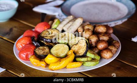 Gegrilltes Gemüse und Würstchen werden auf einem großen weißen Teller serviert, der während eines Sommeressens im Freien eine bunte Auswahl an Farben zeigt. Stockfoto