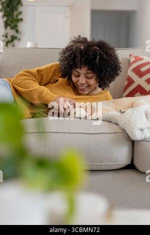 Afroamerikanerin, die sich zu Hause auf dem Sofa liegend und einen Senfpullover trägt, der goldenen Welpen streichelt. Gemütlich, Entspannung, häuslich, Lifestyle, modern, Co Stockfoto