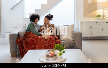 Afroamerikanisches Paar kuschelt unter Überwurfdecke auf grauem Sofa im Wohnzimmer mit Welpen. Gemütlich, modern, liebevoll, entspannt, Zuhause, Innenraum Stockfoto