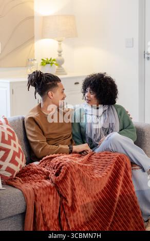 Afroamerikanisches Paar, das auf Sofa mit rot-weißem Kissen und gemütlichem Überwurf im Wohnzimmer sitzt. Gemütlich, intim, Zuhause, Komfort, Entspannung, Umgebungstemperatur, Stockfoto