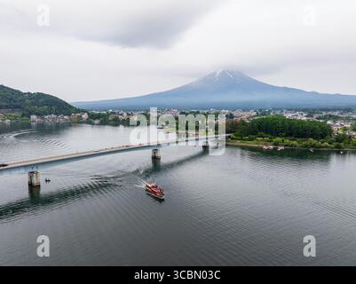 Aus der Vogelperspektive eines Bootes, das auf dem Kawaguchi-See segelt, mit einer Brücke, die sich in Richtung des entfernten, majestätischen, schneebedeckten Mount Fuji, Japan, erstreckt. Stockfoto