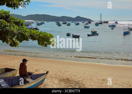 Tropische Strandszene mit Mann, der auf einem Boot mit Fischerbooten und tropischem Laub sitzt, in Buzios, Brasilien Stockfoto