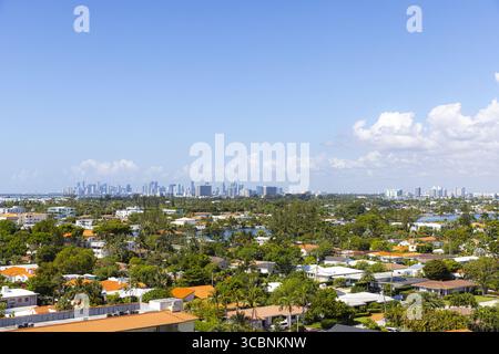 Aus der Vogelperspektive sehen Sie ein dichtes, grünes Baumdach, in dem sich Häuser verstecken, und die ferne Skyline von Miami schimmert unter einem klaren blauen Himmel, Surfside, Florida, USA. Stockfoto