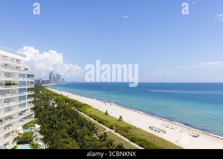 Aus der Vogelperspektive sehen Sie weiße Gebäude, die hoch über dem azurblauen Himmel stehen und auf den türkisfarbenen Atlantik treffen, einen Küstentanz aus moderner Architektur und natürlicher Schönheit, Surfside, Florida, USA. Stockfoto