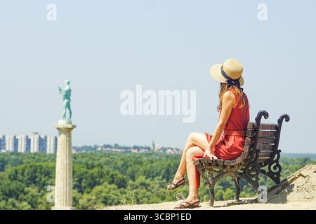 Frau in Kleid und Hut genießt einen Sommertag in Kalemegdan, Serbien. Das Pobednik-Denkmal und eine Reisende auf einer Bank in der Belgrader Festung Stockfoto