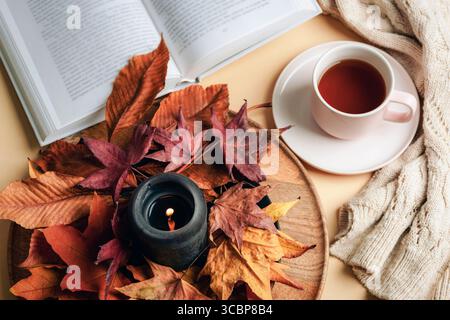 Tasse Tee mit Buch, schwarzer Kerze und Herbstlaub auf Holzteller mit warmem Pullover. Gemütliche Herbststimmung und Entspannungskonzept. Draufsicht, flach. Stockfoto