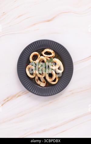 Tintenfischringe, garniert mit Kräutern, in kreisförmigem Muster auf einer dunklen Platte über einer hellen Marmoroberfläche angeordnet. M+S Meeresfrüchte von oben fotografiert. Stockfoto