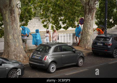 Erhöhte Sicht auf Menschen, die bei regnerischem Wetter auf einer von Bäumen gesäumten Straße laufen und stehen, einige tragen blaue Ponchos, und geparkte Autos sind sichtbar. Stockfoto