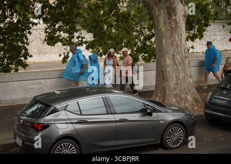 Erhöhte Sicht auf Menschen, die bei regnerischem Wetter auf einer von Bäumen gesäumten Straße laufen und stehen, einige tragen blaue Ponchos oder Regenmäntel. Stockfoto