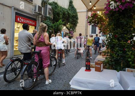 Touristen mit dem Fahrrad fahren an einem Sommertag entlang einer engen kopfsteingepflasterten Straße, die von Geschäften und Cafés gesäumt ist. Via della Vite in Rom, Italien. Stockfoto