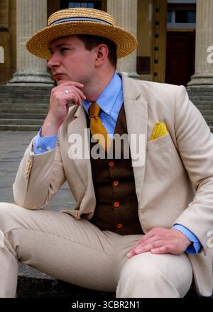 Ein Mann in einem stilvollen Outfit, einschließlich Strohhut, Anzug, Weste und Krawatte, sitzt und berührt nachdenklich sein Kinn. Stockfoto
