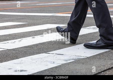 Japanischer Angestellter mit flachem Winkel, der die Fußgängerzone in städtischer Umgebung überquert. Der Stil besteht aus Japans lässiger, sommerlicher Business-Kleidung. Stockfoto