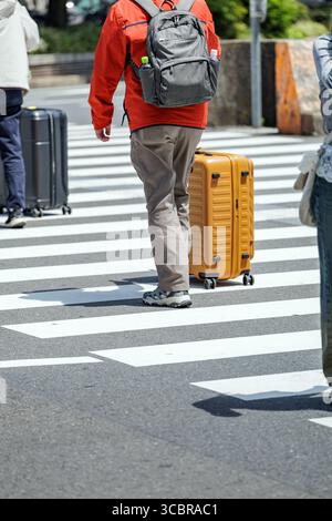 Touristen, die mit Gepäck und Rucksäcken eine urbane Fußgängerzone überqueren, zeigen urbane Reisen und Mobilität in einer urbanen Umgebung. Stockfoto