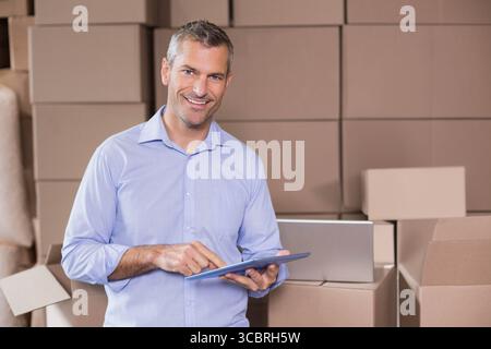 Mann im mittleren Erwachsenenalter, der das Tablet in der Nähe des Laptops anklopft, während er im Lager steht und von Kartons umgeben ist Stockfoto