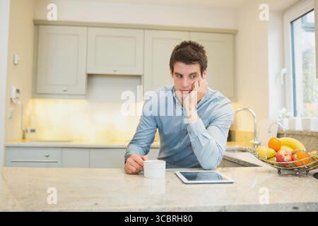 Mann, der den Ellbogen auf der Marmorarbeitsfläche in der Küche ruht, während er den Becher neben dem Tablet packt Stockfoto