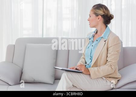 Frau in Geschäftskleidung schreibt mit Stift in spiralgebundenem Notizbuch auf Sofa zu Hause, Kopierraum Stockfoto