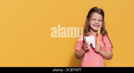 Ein glückliches Mädchen hält eine Zahnbürste und einen Plastikzahn in den Händen. Eine isolierte Panoramaaufnahme. Ein Ort für Ihre SMS. Kariesprophylaxe im Kindesalter. Stockfoto