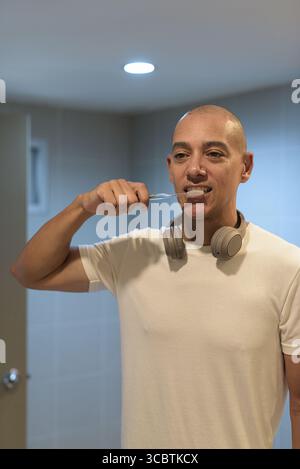 Kahler hispanischer Mann in lässigem weißem T-Shirt mit Kopfhörern, Zähneputzen im modernen Badezimmer nach dem Fitnesstraining. Körperhygiene und gesunder Lebensstil Stockfoto