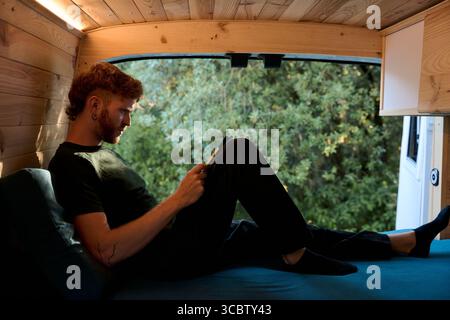 Junger Mann, der sich auf dem Bett entspannt und im Wohnmobil eine Tablette liest Stockfoto