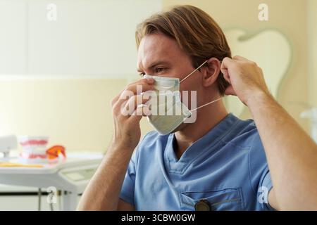 Kaukasischer junger erwachsener Mann, der medizinische Peelings trägt, die chirurgische Maske auf dem Gesicht in der klinischen Umgebung, Vorbereitung für das Gesundheitswesen Eingriff, fokussierter Ausdruck sichtbar Stockfoto