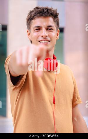 Ein lächelnder junger Mann, der auf die Kamera zeigt und draußen rote Kopfhörer trägt. Vertikal Stockfoto