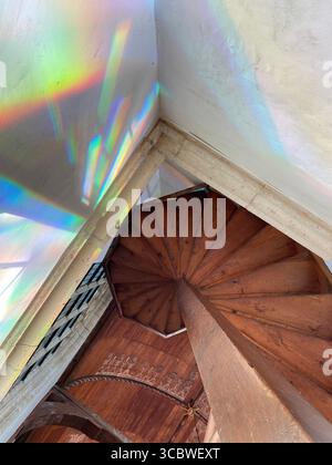 Unkonventioneller Lichtwinkel, der sich in geometrisch unterschiedlichen Räumen von einem Buntglasfenster in einer Kirche auf einer Holztreppe bricht Stockfoto
