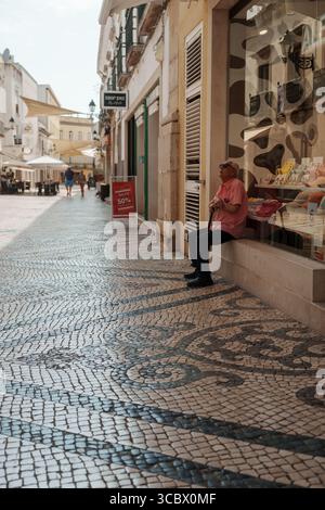 Algarve, Portugal – 2025.06.28 ein älterer Mann in einem rosa Hemd sitzt auf einem Läden-Felsvorsprung und ruht im Schatten einer mit Mosaiken gekachelten Einkaufsstraße im h Stockfoto